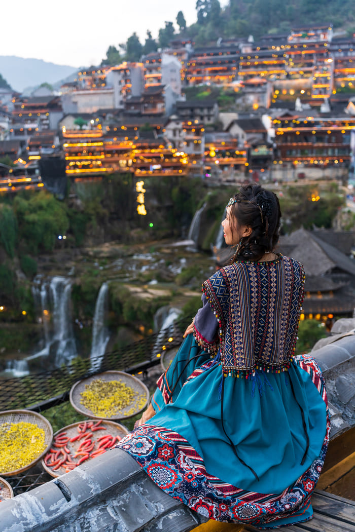 Una mujer vestida con ropa tradicional colorida mira hacia la ciudad antigua de Lijiang, China, mientras se iluminan las linternas al atardecer sobre una cascada.