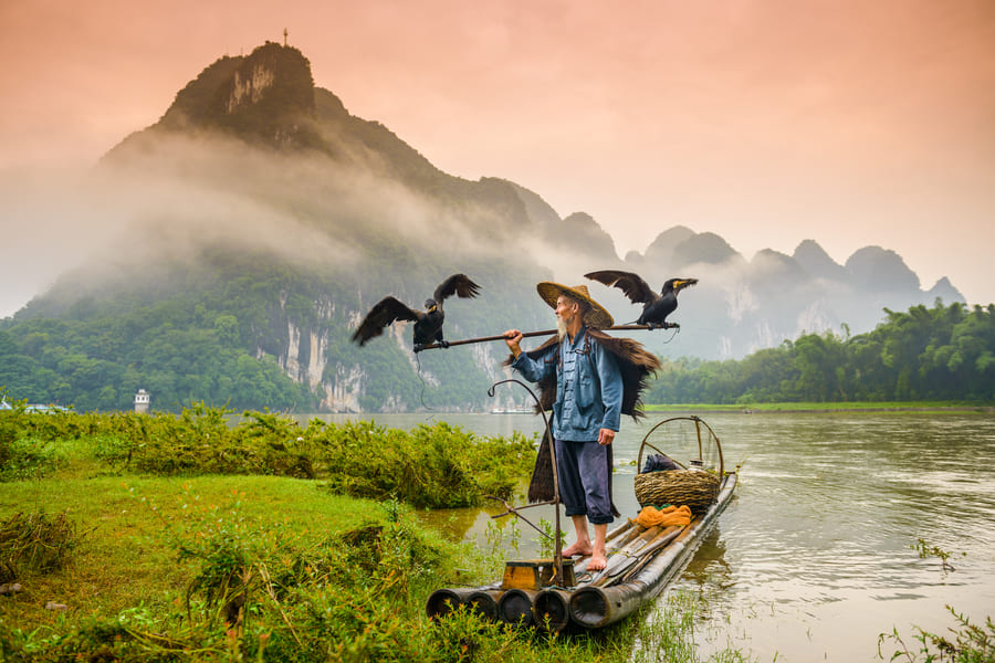 Pescador de cormorán sobre una canoa en el río Yulong en Yansgshuo con montañas de fondo